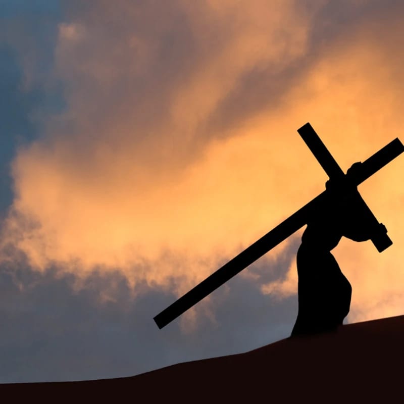 A silhouette of Jesus Christ carrying a large wooden cross on his back up a hill against a dramatic sunset sky with orange and dark blue clouds.